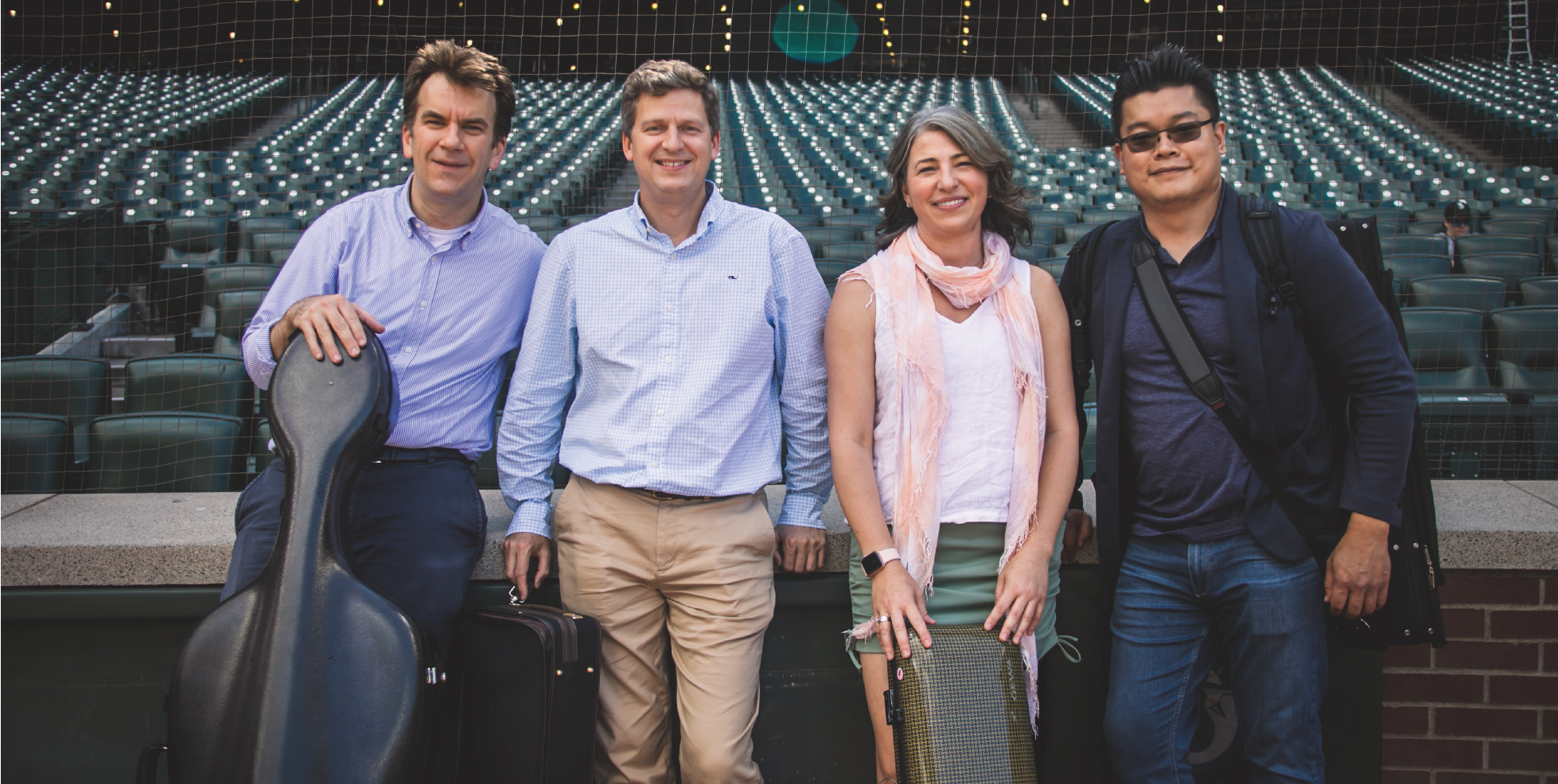 Four musicians, each holding an instrument case, standing in front of stadium seating.